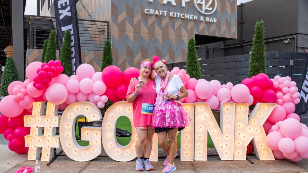 Two participants pose with medals at the #GoPink display outside Tap 42 after the Partners Pink Run Fort Myers 2025.