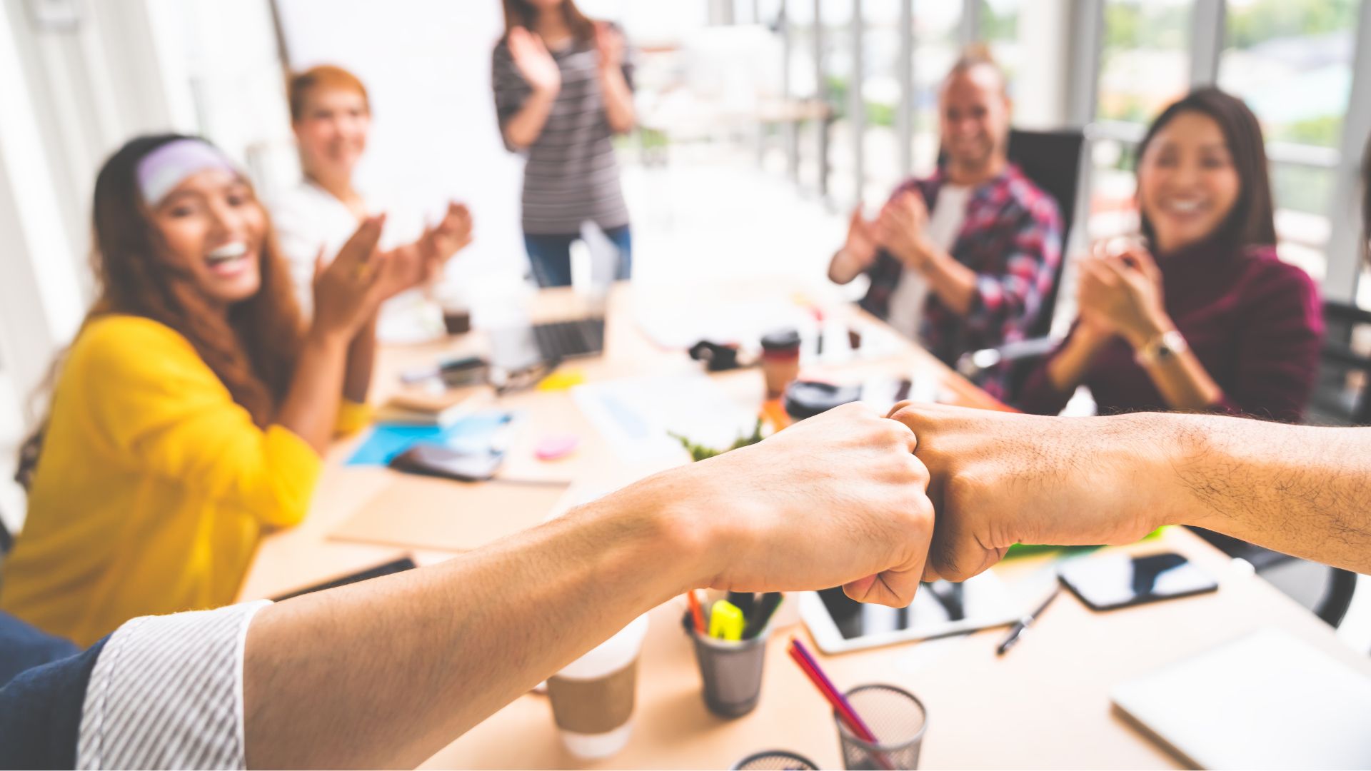 Two team members fist bump across a meeting table while others smile and applaud, symbolizing trust, collaboration, and shared success.