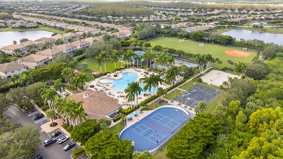 Aerial close-up of Bella Terra amenities in Estero, FL, featuring the Tuscan-style clubhouse, resort-style swimming pool, tennis courts, basketball court, playground, and lakefront setting with homes for sale nearby.