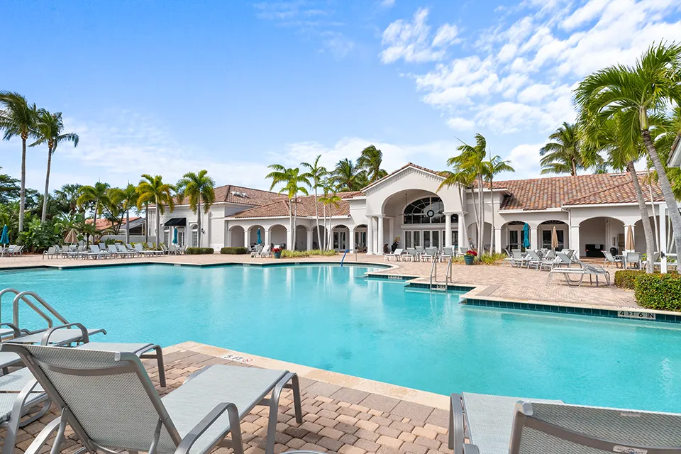Resort-style swimming pool and clubhouse at Cascades at Estero 55+ in Estero, FL, featuring lounge seating, umbrellas, palm trees, and patio with active adult homes for sale nearby.