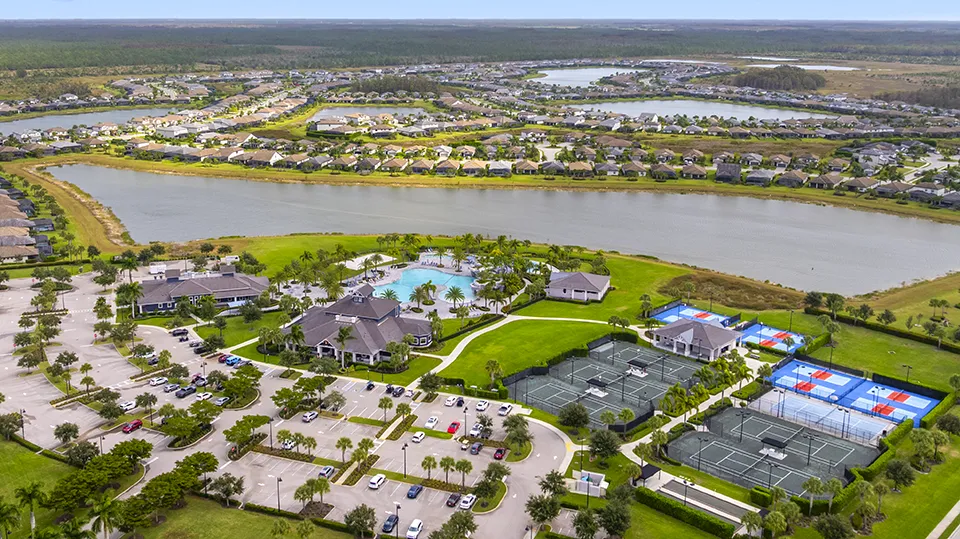 Aerial close-up of The Place at Corkscrew amenities in Estero, FL, featuring the two-story clubhouse, resort-style pools with waterslide, tennis and pickleball courts, basketball court, and lakefront setting with homes for sale nearby.