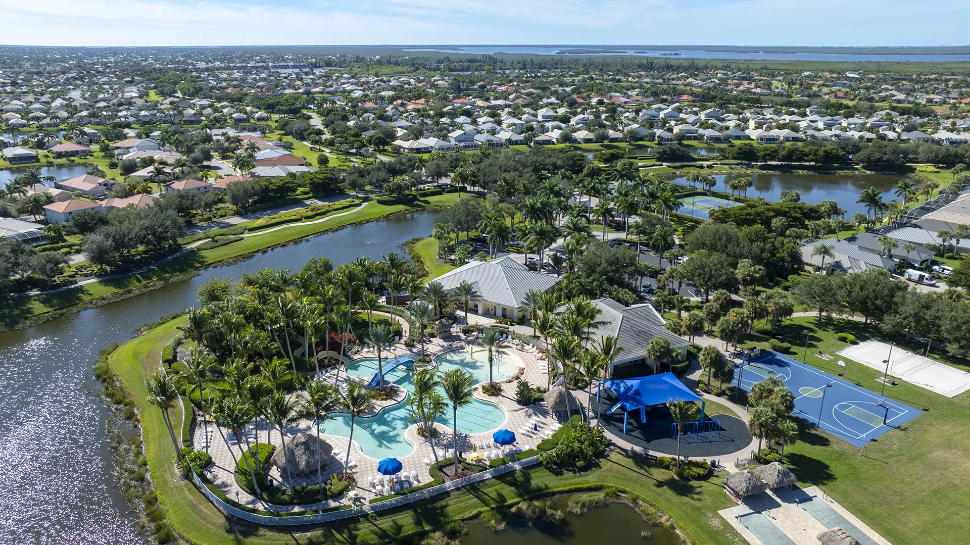Aerial view of Sandoval in Cape Coral, FL, showing the resort-style clubhouse, lagoon swimming pool with waterslide, tennis and pickleball courts, basketball court, playground, lake views, and new homes for sale.