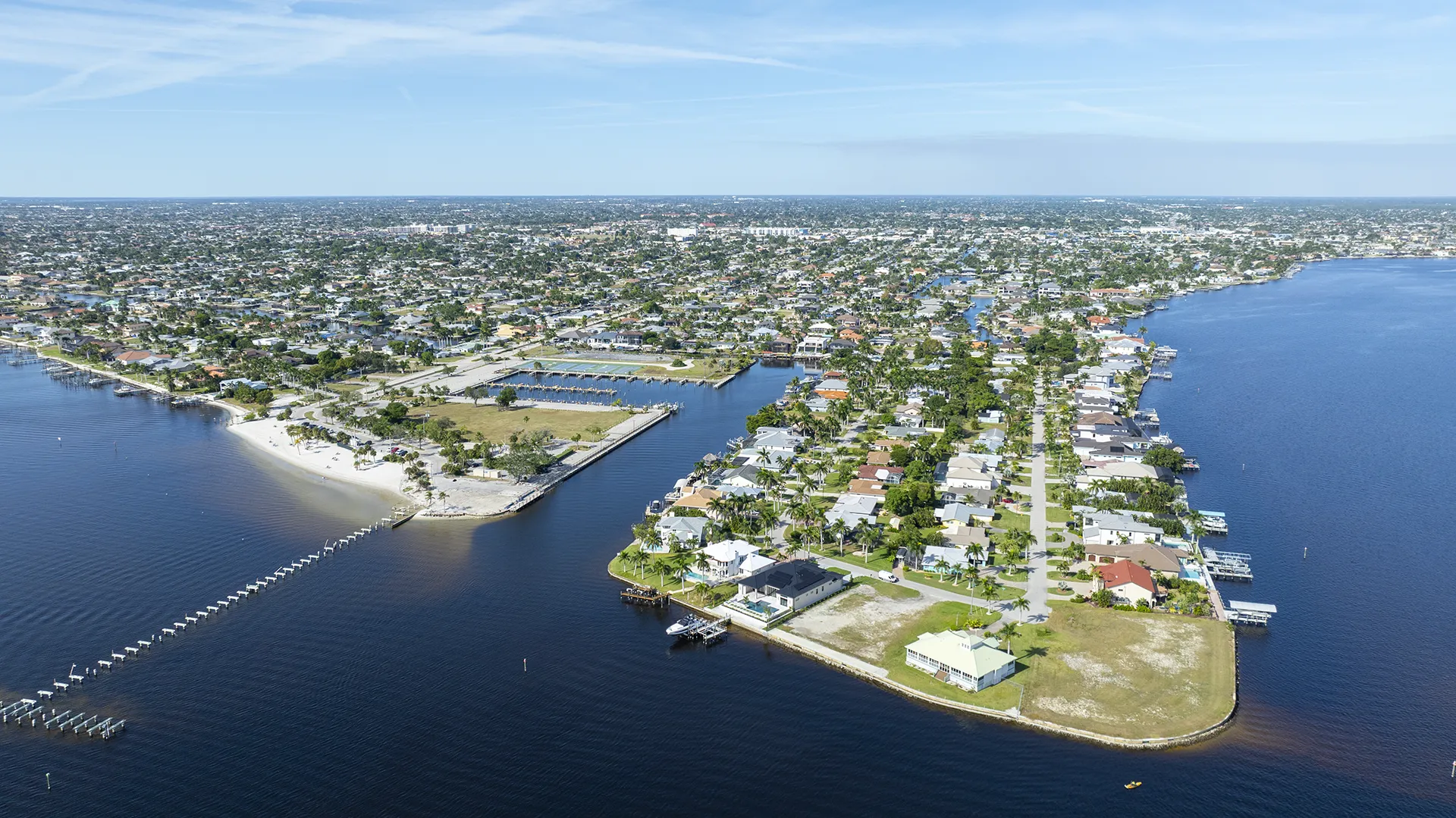 Aerial view of Cape Coral Yacht Club area in Cape Coral, FL, featuring the public beach, resort-style pool, tennis courts, boating ramp and slips, community center, and surrounding waterfront homes for sale with direct river access.