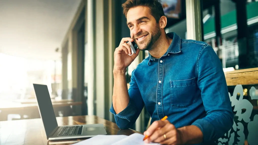 Real estate agent on a phone call at a coffee shop, smiling and taking notes — building trust in real estate means becoming the agent people actually call