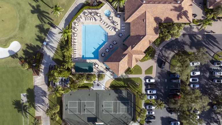 Aerial view of a clubhouse with pool, tennis courts, parking, and adjacent golf course